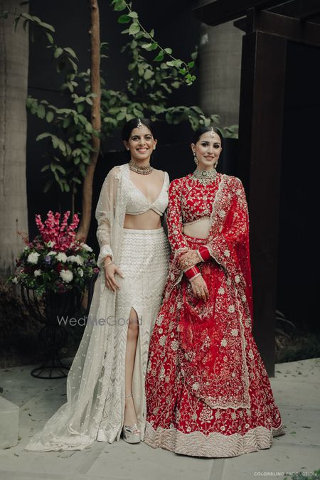 Photo of Wedding day capture of the bride with her sister in a red lehenga and the sister of the bride in a high slit white lehenga