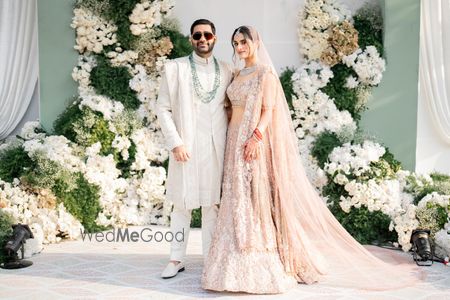 Photo of Couple portrait with gorgeous floral backdrop with the bride in a pastel peach lehenga and the groom in an all-white sherwani