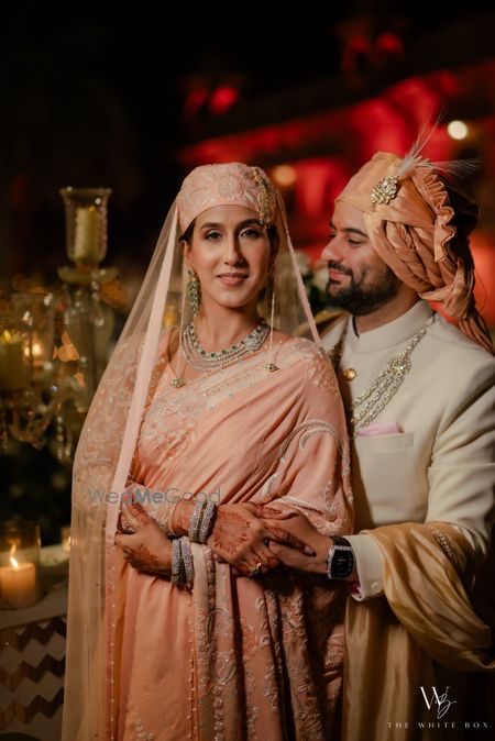 Photo of Lovely couple shot after getting married with the bride in a peach saree with headgear and the groom in a co-ordinated peach safa and ivory sherwani