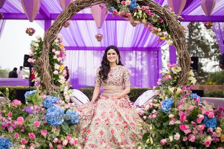 Photo of A bride sitting under a wreath of flowers in a nude peach lehenga for her mehendi.